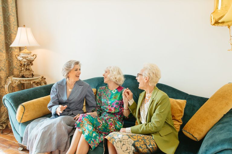 Three older women laughing together on a teal couch